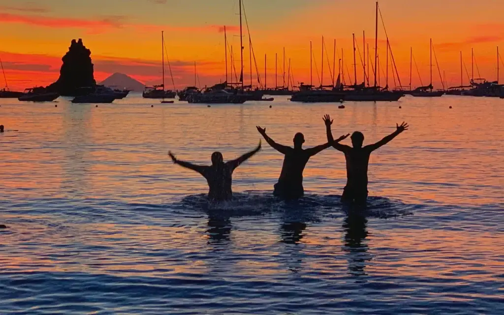 Tramonto alle Eolie con tre amici in acqua tra barche a vela e faraglioni – spirito festivo e comunitario del Festival EOLIÈ.
